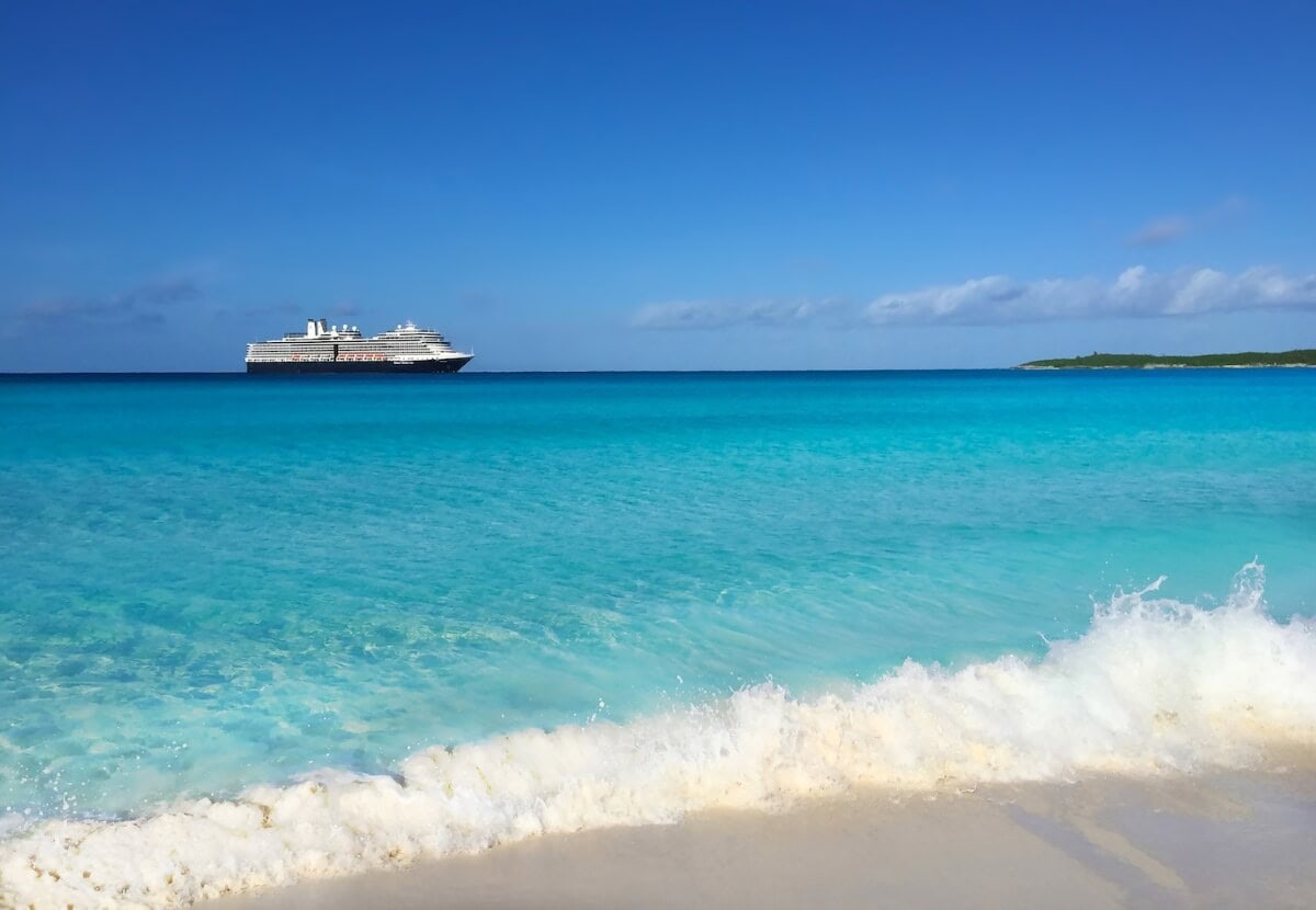 A cruise ship sails on a calm turquoise sea near a sandy beach under a clear blue sky.