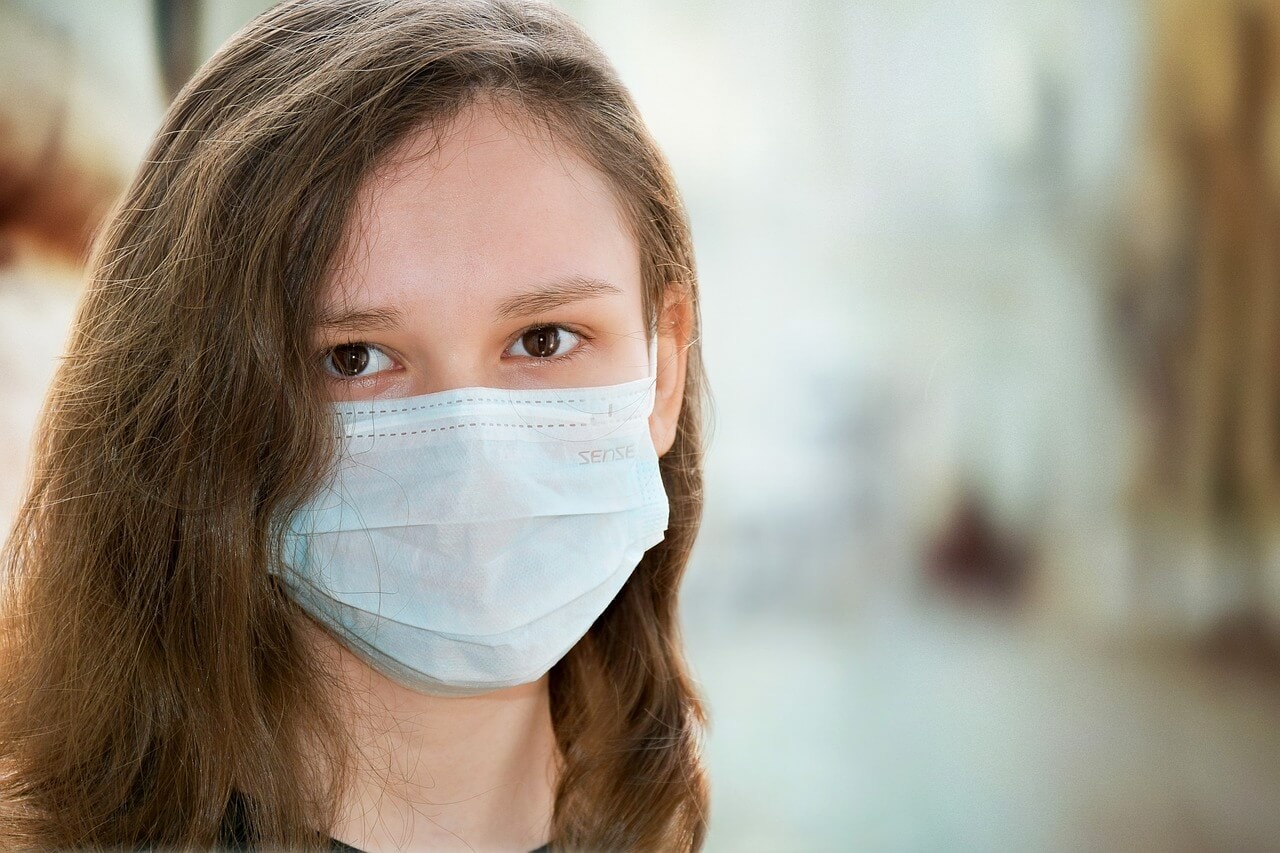 A young person with long hair wearing a blue surgical mask, looking directly at the camera. The background is blurred, reminiscent of a serene scene you might want to protect with cruise travel insurance.