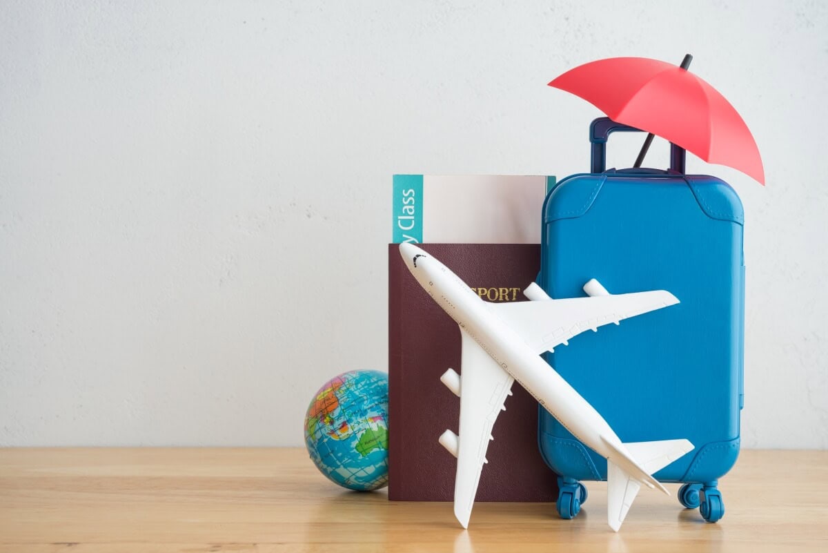 A small blue suitcase, passport, boarding pass, toy airplane, miniature globe, and red umbrella are displayed on a wooden surface against a plain background, all set for cruise travel adventures.