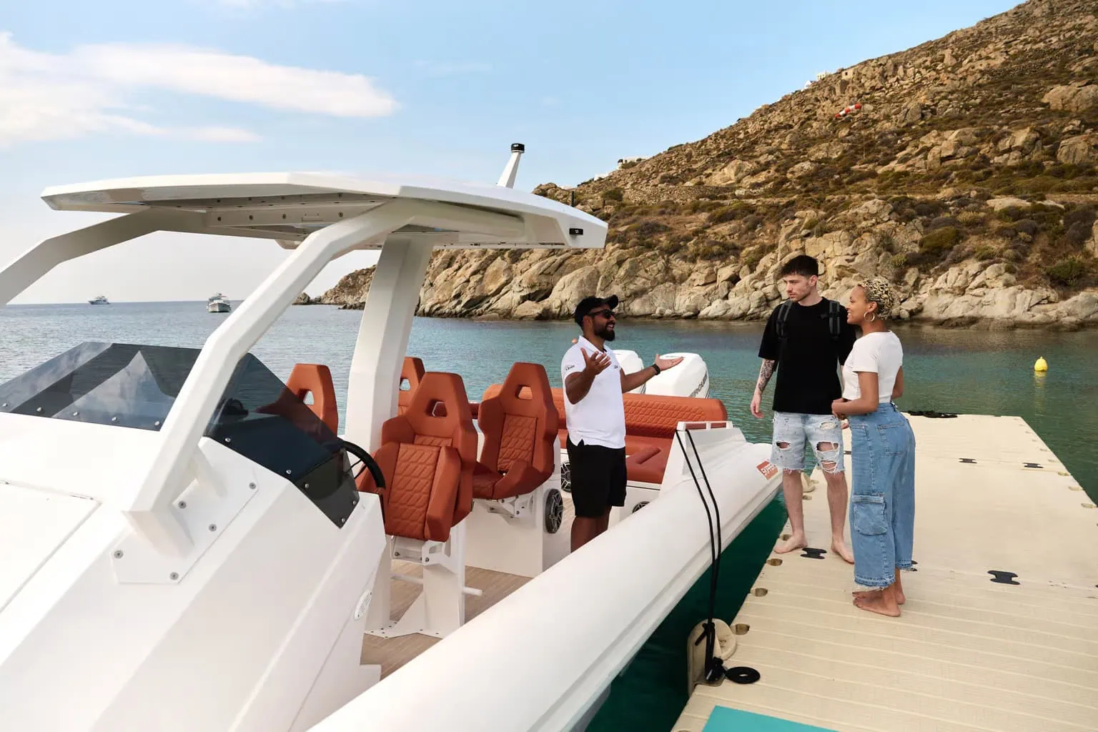 Three people stand on a dock next to a white boat with red seats, in front of rocky terrain and blue water. One person, wearing a hat, seems to be explaining something to the other two, as if preparing them for an upcoming tour on their European cruise.