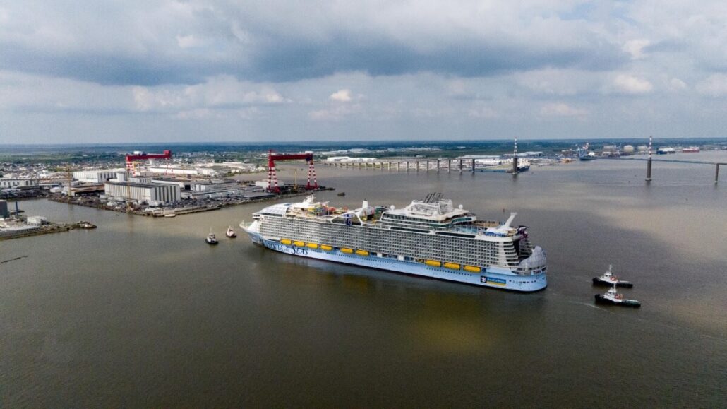 Aerial view of the Utopia of the Seas embarking from a port, surrounded by tugboats, with a cityscape and bridge in the background.