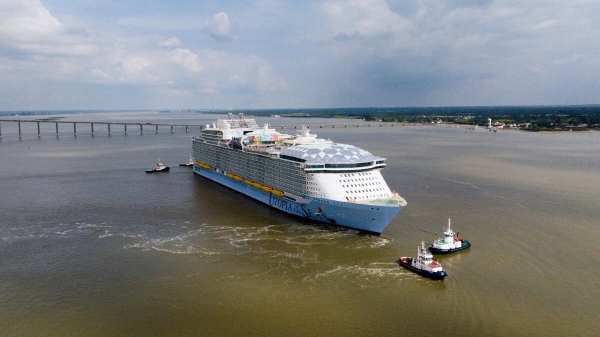 Aerial view of the Utopia of the Seas, a large cruise ship, being escorted by two tugboats on a wide river, with a long bridge visible in the background.