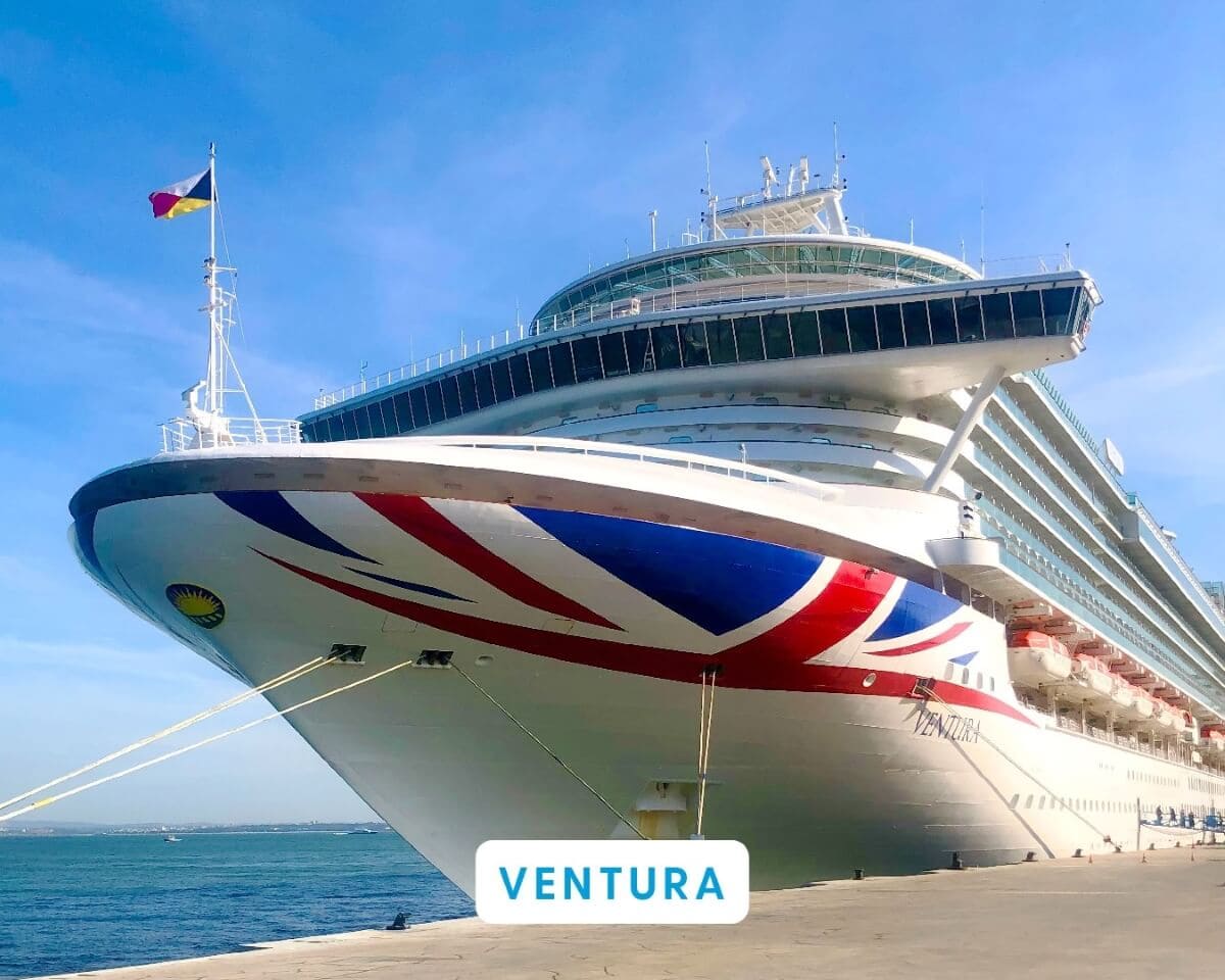 A large P&O Cruise ship named Ventura is docked at a port, showcasing the British flag's colors alongside a Romanian flag hoisted on its mast. The sky is clear and blue, highlighting the vessel's grandeur and significant size.