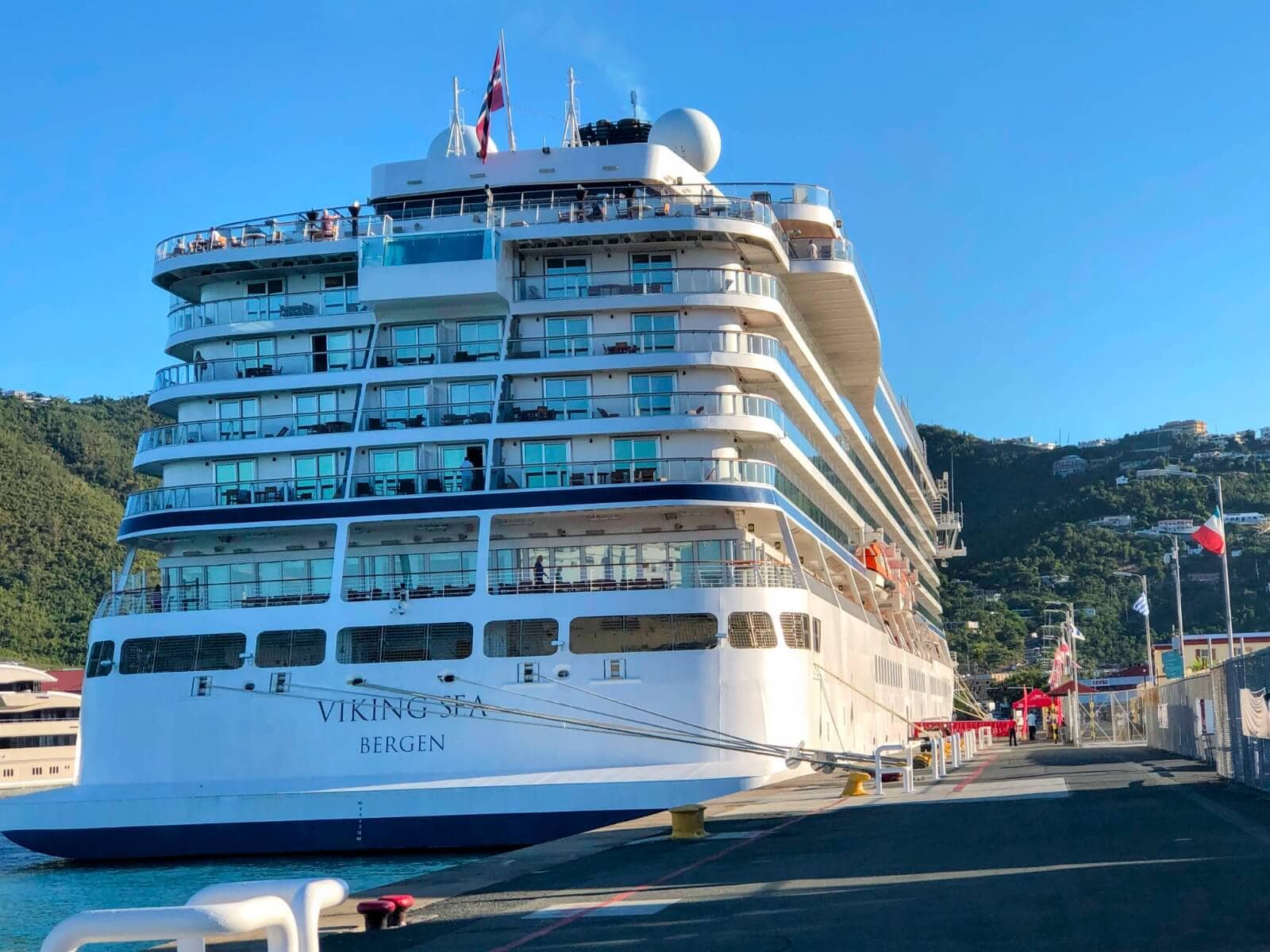 A large Viking Cruise Ship, the Viking Sea, docked at a port with a clear blue sky and hilly landscape in the background, showcasing its impressive size among the fleet.