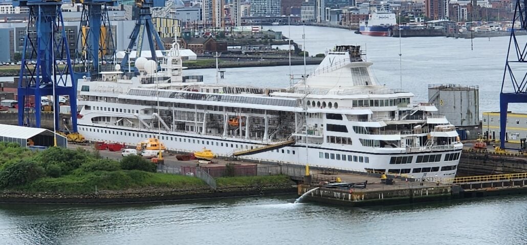 A large white cruise ship, preparing for its 300-day round-the-world cruise, is docked for maintenance in a shipyard. It’s surrounded by cranes and industrial structures with a cityscape in the background.