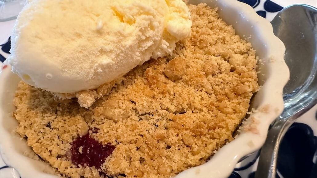 A white ceramic dish of berry crumble topped with a scoop of vanilla ice cream, reminiscent of sweet indulgences enjoyed during an Alaska trip, with a silver spoon beside it.